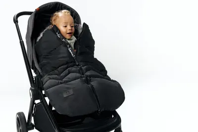 A baby wrapped in a black footmuff is sitting in a stroller against a white background.