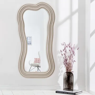 A decorative mirror with a wavy frame is mounted on a white wall. A vase with dried flowers sits on a stack of books in the foreground.