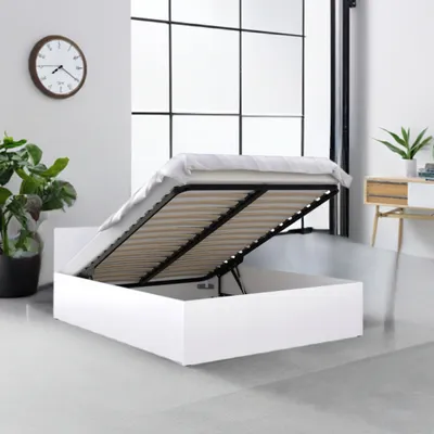 A modern white bed with its storage compartment open, revealing the slatted base, in a well-lit room with a large window and a clock on the wall.