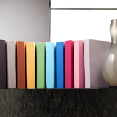 A row of colorful fitted sheets are displayed on a dark wooden surface in front of a light gray wall and a bright window.