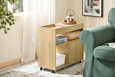 A wooden rolling cart with storage compartments is placed next to a green armchair in a room with natural light coming from a window.