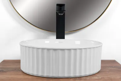 A white ceramic washbasin with a fluted exterior sits on a wooden surface in front of a large round mirror and a black faucet.