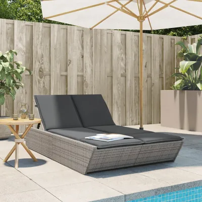 A double sun lounger with dark gray cushions sits on a patio next to a swimming pool, with a wooden fence and plants in the background.