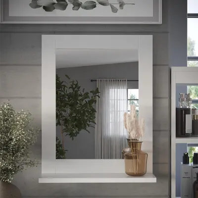 A white framed mirror with a shelf below it, placed against a gray textured wall. A plant is visible to the left, and a vase with dried flowers is on the shelf.