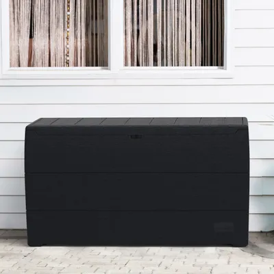 A black storage box sits on a patio in front of a white wall with two windows.