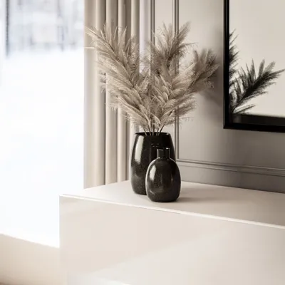 A close-up shot of two dark vases with dried pampas grass in one, placed on a white glossy surface, with a blurred background of curtains and a mirror reflection.