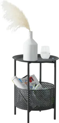 A black metal side table with a white vase and dried flowers, and a glass of water on top, and magazines in the basket below.