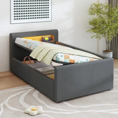 A grey upholstered bed with its mattress lifted to reveal storage compartments underneath. The headboard has integrated LED lighting and a small shelf. A plant and artwork are visible in the background.