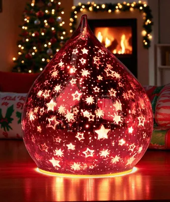 A red glass teardrop-shaped lamp with star cutouts is lit up, placed on a wooden surface in a festive, Christmas-themed room with a decorated tree and a fireplace in the background.