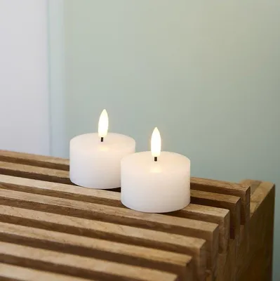 Two white LED tea lights with illuminated flames sit on a slatted wooden surface against a pale green and white background.