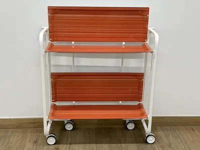 A white metal rolling cart with two orange shelves is shown against a white wall and wooden floor.