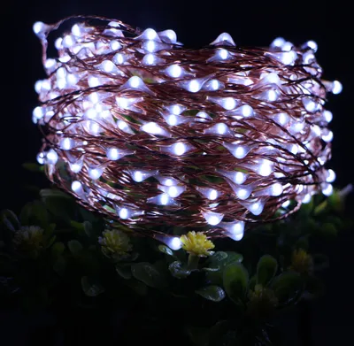 A close-up of a coiled string of white LED lights on a copper wire, resting on green foliage with small yellow flowers.