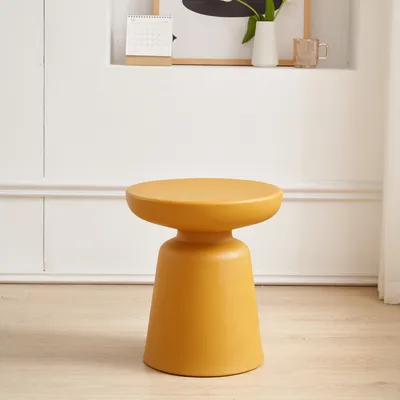 A yellow side table is placed on a light wooden floor in front of a white paneled wall. A shelf above the table holds a calendar, a vase with flowers, and a glass mug.