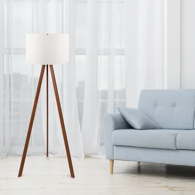 A floor lamp with a white shade and wooden tripod base stands next to a light blue sofa in a room with sheer white curtains.