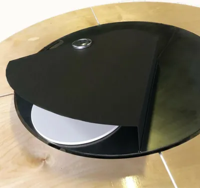 A close-up view of a black pizza lid partially covering a white pizza stone, set on a light wood table.
