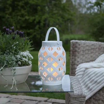 A white lattice lantern with a warm glow sits on a glass table next to a potted plant and a wicker chair with a blanket.