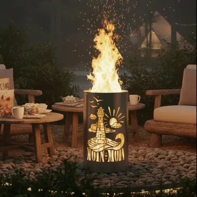 A fire pit with a lighthouse design is lit at night in a garden setting, with chairs and tables nearby.