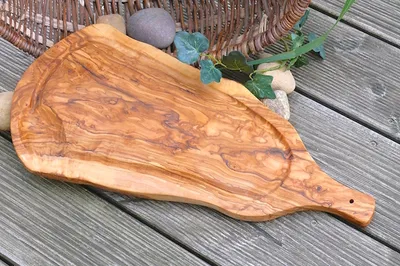 A rustic olive wood cutting board with a juice groove is placed on a wooden deck, with a wicker basket and stones in the background.