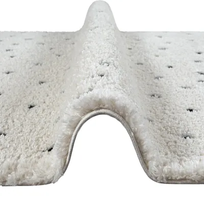 A close-up, angled view of a beige rug with black dots, showing its high pile texture and a rolled edge.