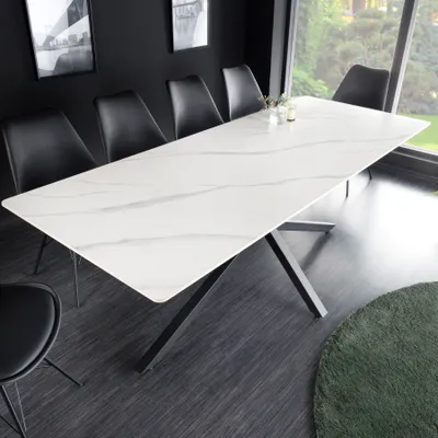 A modern dining table with a white ceramic top and a black metal base, set in a dining room with black chairs and artwork on the wall.