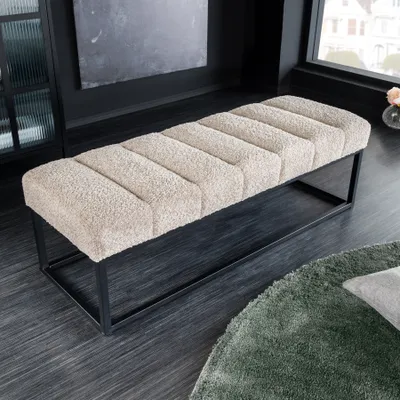 A bouclé bench with a black metal frame is placed on a dark wooden floor next to a window. A green rug is visible in the foreground.