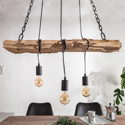 A rustic chandelier made from a piece of driftwood with three exposed Edison-style bulbs hanging from black cords, suspended by chains. It is shown above a wooden dining table with black chairs in the background.