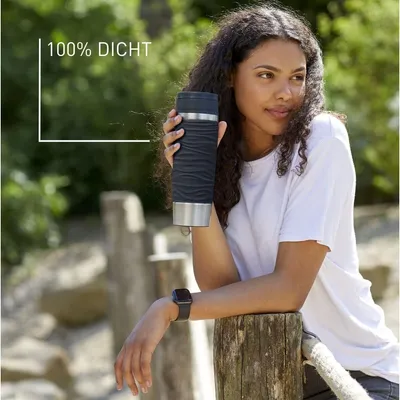 A woman is holding a black travel mug outdoors, leaning on a wooden post.