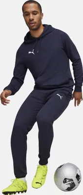 A man wearing a blue Puma tracksuit and yellow soccer cleats poses next to a soccer ball.