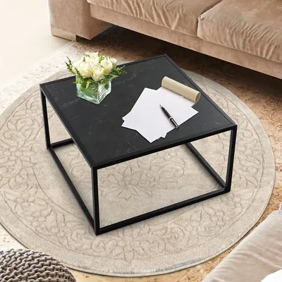 A black marble-look coffee table with a metal frame sits on a circular rug in a living room setting, next to a beige sofa. There are papers and a pen on the table, along with a vase of white flowers.