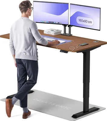 A man stands at a height-adjustable desk with two monitors, using a mouse. A mat is on the floor in front of the desk.