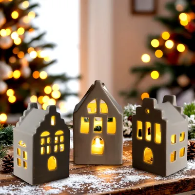 Three ceramic house-shaped candle holders are displayed on a wooden surface dusted with artificial snow, with a blurred Christmas tree and lights in the background.