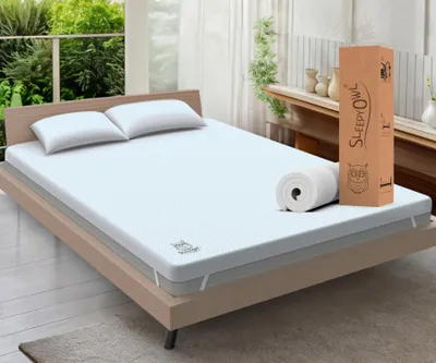 A mattress topper is displayed on a bed with two pillows, next to its packaging and a rolled-up topper.