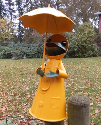 A garden decoration of a frog wearing a yellow raincoat and holding an umbrella, standing on a wooden post in a garden setting with fallen leaves.