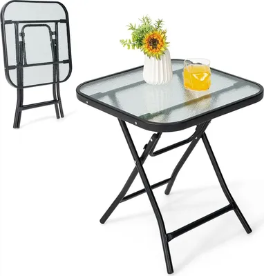 A square glass-top folding table with a black metal frame, shown with a vase of flowers and a glass of orange juice on top. A second, folded table is visible in the background.