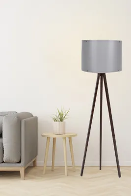 A floor lamp with a gray shade and a wooden tripod base stands next to a small wooden side table with a potted plant on it, and a portion of a gray sofa is visible on the left.