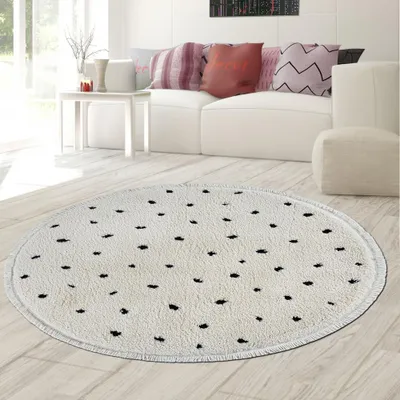 A round, high-pile rug with a black polka dot pattern is placed on a light wooden floor in a living room setting. A white sofa with decorative pillows is visible in the background, along with a white side table and a large window.