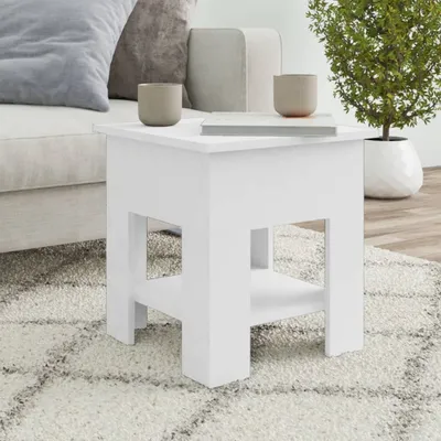 A white side table is placed on a rug next to a sofa, with two decorative cups and a book on top. A potted plant is visible in the background.
