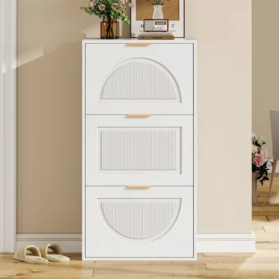 A white shoe cabinet with three flip-down doors is shown in a room with light beige walls and a wooden floor. A pair of slippers is on the floor in front of the cabinet.