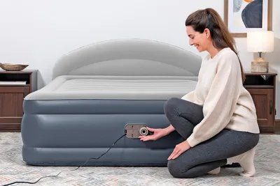 A woman is interacting with an air mattress in a bedroom setting. There are nightstands on either side of the bed, one with a lamp and artwork on the wall above.