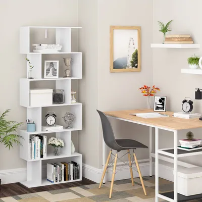 A white bookshelf with 6 compartments is filled with books and decorative items, placed in a room next to a desk with a chair.
