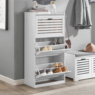A white shoe cabinet with multiple shelves and flip-down compartments is shown in a room with gray walls and a wooden floor. A gray coat is hanging on the wall, and a brown hat is placed on a nearby bench.