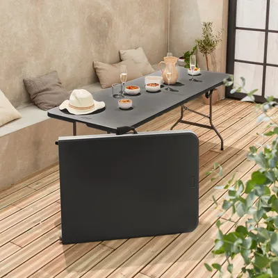 A folded dark gray party table is in the foreground, with an unfolded dark gray party table set up on a wooden deck in the background. The table is set with glasses, bowls of food, and a hat.