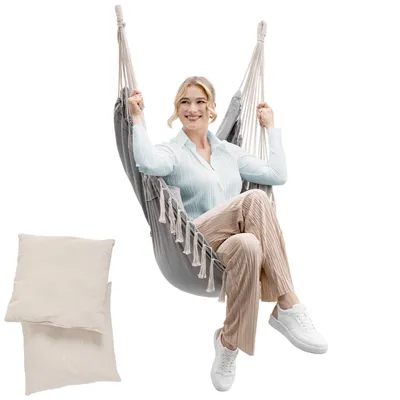 A woman sits in a hanging chair with two pillows beside her.