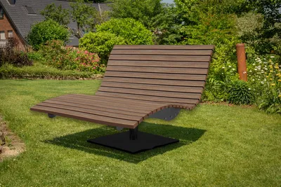 A wooden chaise lounge sits on a green lawn in a garden setting, with a house and trees in the background.