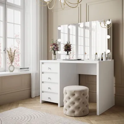A white vanity table with a mirror and lights, a tufted stool, and decorative flowers in vases, set in a well-lit room with a large window.