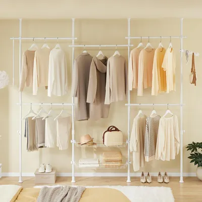 A white telescoping garment rack with multiple shelves and hanging clothes, set up in a room with a bed in the foreground.