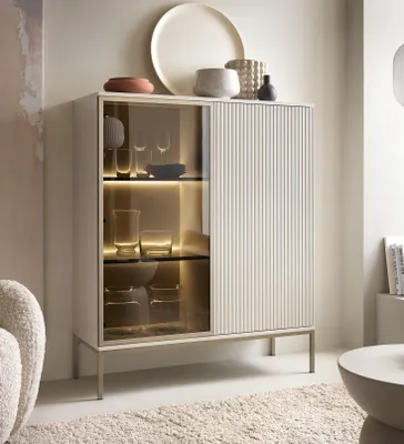 A modern display cabinet with glass doors and fluted panels, showcasing glassware and decorative items. The cabinet is placed in a well-lit, stylish room with a textured rug and a plush armchair.