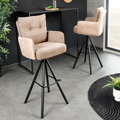 Two beige bar stools with black legs are placed in a modern interior setting, with a black bar table in the background and a decorative rug on the floor.