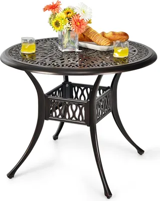 A round patio table with a decorative top and a basket-like base, set against a white background. It is adorned with a vase of flowers, two glasses of yellow liquid, and a plate of bread.