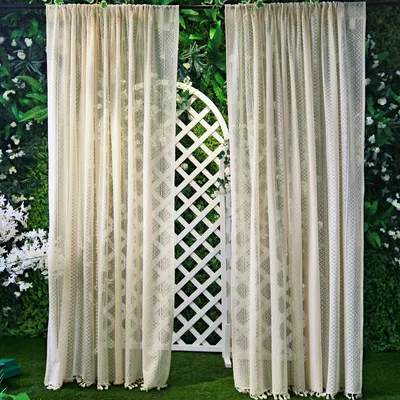 Two cream-colored lace curtains are hung in front of a white lattice archway, with lush green foliage and white flowers in the background.
